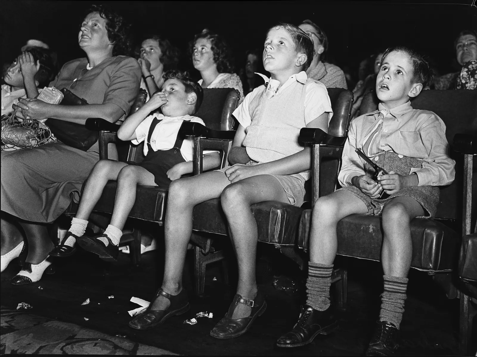 In the stalls of a picture theatre a mother with a string bag and three boys in shorts and long socks stare up at the screen; the smallest has his thumb in his mouth, the boy on the right holds a toy pistol, and sweet papers lie on the floor.