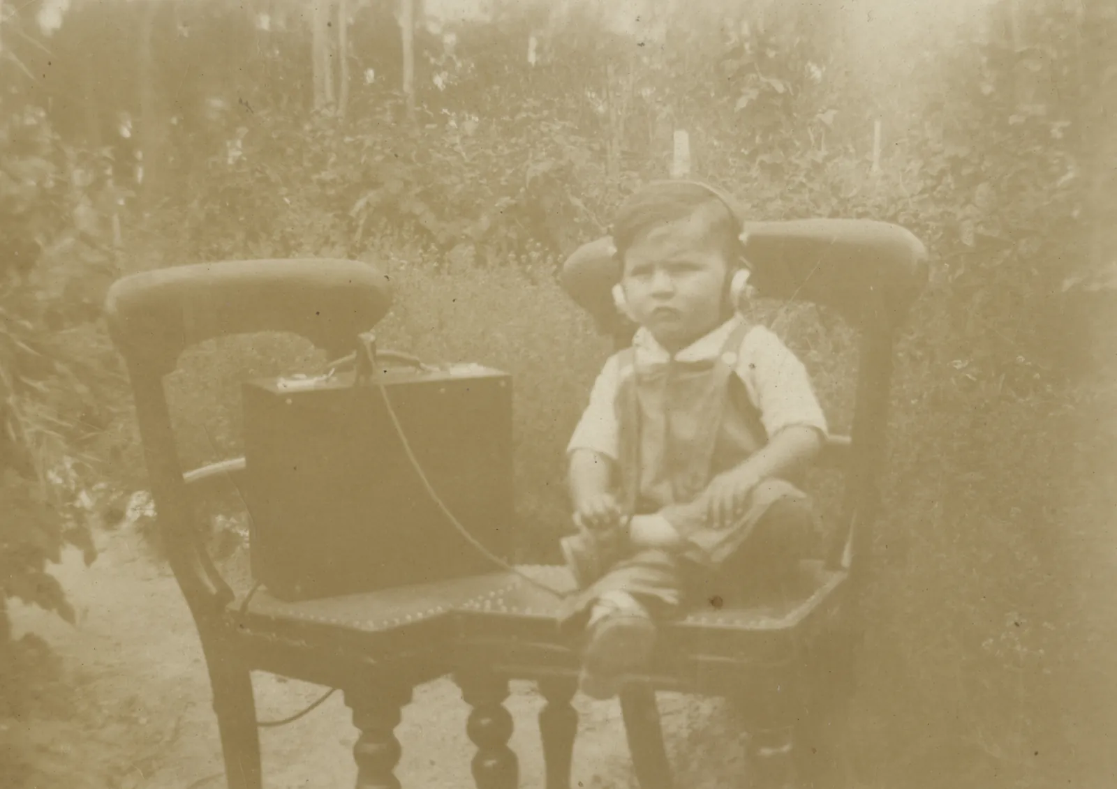 A small boy in a shirt and overalls sits scowling on a big carved chair in a garden, wearing headphones wired to a box wireless set on the seat beside him.