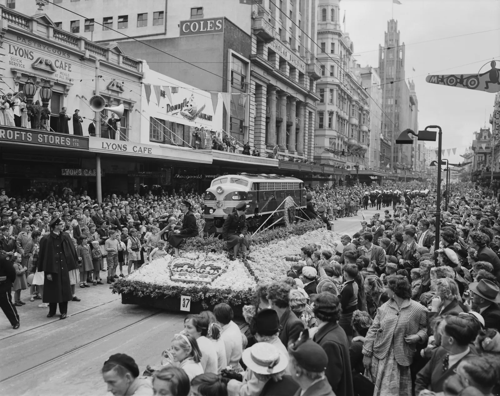 Swanston Street packed from the kerb to the shop verandas as a float of flowers carrying a model diesel locomotive passes behind a band; Moomba’s banners and bunting hang over the street, and the Manchester Unity building rises at the end of it.