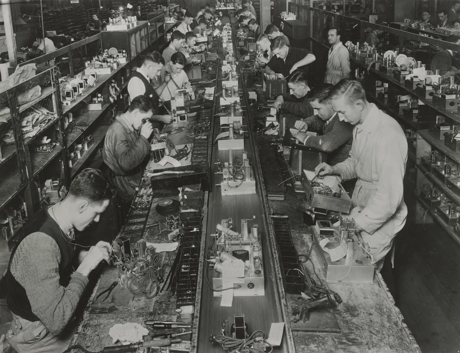 Looking down a long double workbench in a radio factory: rows of young men in shirtsleeves and cardigans wire and solder radio chassis, with shelves of parts on either side and a foreman in a white coat at the back.